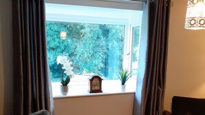 A living room with a sofa, a table, a window with curtains, a clock, and a plant.