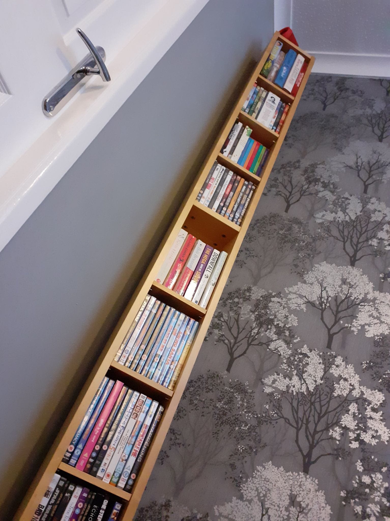 A bookshelf filled with books and DVDs against a wall with a tree pattern carpet.
