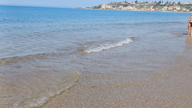 A beach with clear water, pebbles, and a distant town. - Montereale Holiday Home (Realmonte)