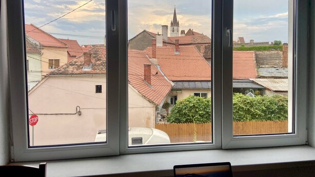 Visão pela janela dividida em três painéis, mostrando uma área residencial com diversos edifícios, incluindo uma igreja com torre e uma placa de pare na esquerda.
