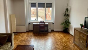 Uma sala de estar com piso de madeira em padrão herringbone, um sofá, uma mesa de centro de madeira, um radiador, uma janela com persianas, uma escrivaninha, uma cadeira, uma planta em vaso e uma televisão sobre um armário de madeira.