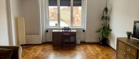 A living room with a herringbone wood floor, a sofa, a wooden coffee table, a radiator, a window with blinds, a desk, a chair, a potted plant, and a TV on a wooden cabinet.