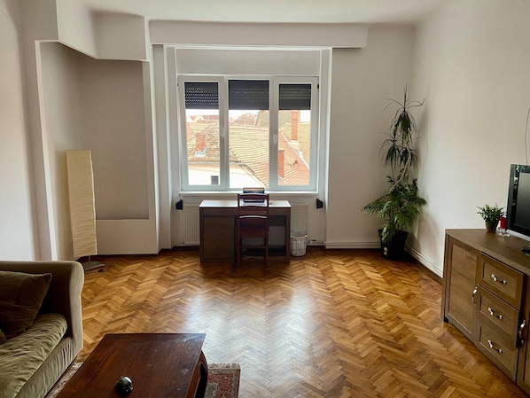 A living room with a herringbone wood floor, a sofa, a wooden coffee table, a radiator, a window with blinds, a desk, a chair, a potted plant, and a TV on a wooden cabinet.