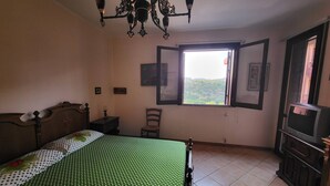 A bedroom with a bed, a wooden dresser, a chair, a window with a view, and a chandelier. - Belvedere House Noto Alta (Noto)