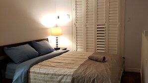 Une chambre à coucher avec un lit, des oreillers, une table de chevet avec une lampe et une fenêtre avec des stores.
