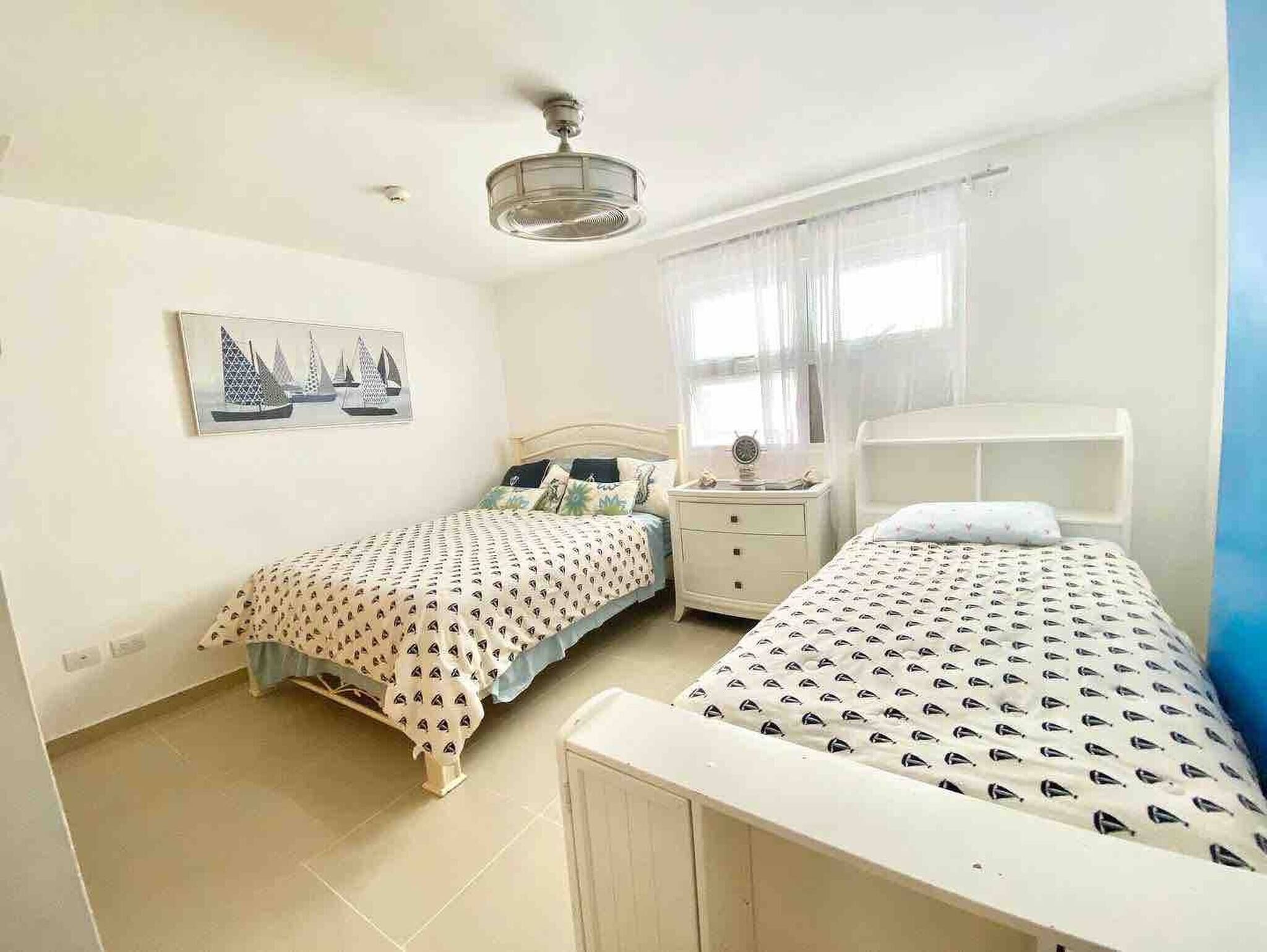 A bedroom with two single beds, a nightstand, a ceiling fan, a window with sheer curtains, and a wall-mounted picture.