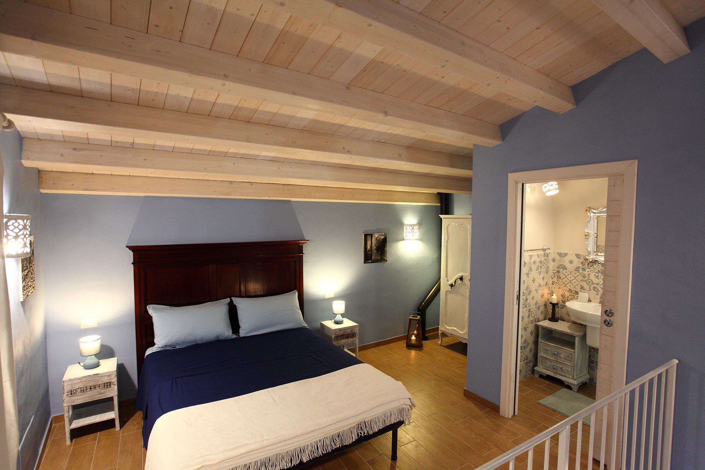 A bedroom with a wooden ceiling, a bed with a blue bedspread, bedside lamps, a nightstand, and a view of a bathroom with a sink and mirror.
