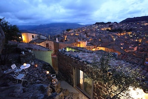 A stone building with a glass door, outdoor seating, and a view of a town at dusk.