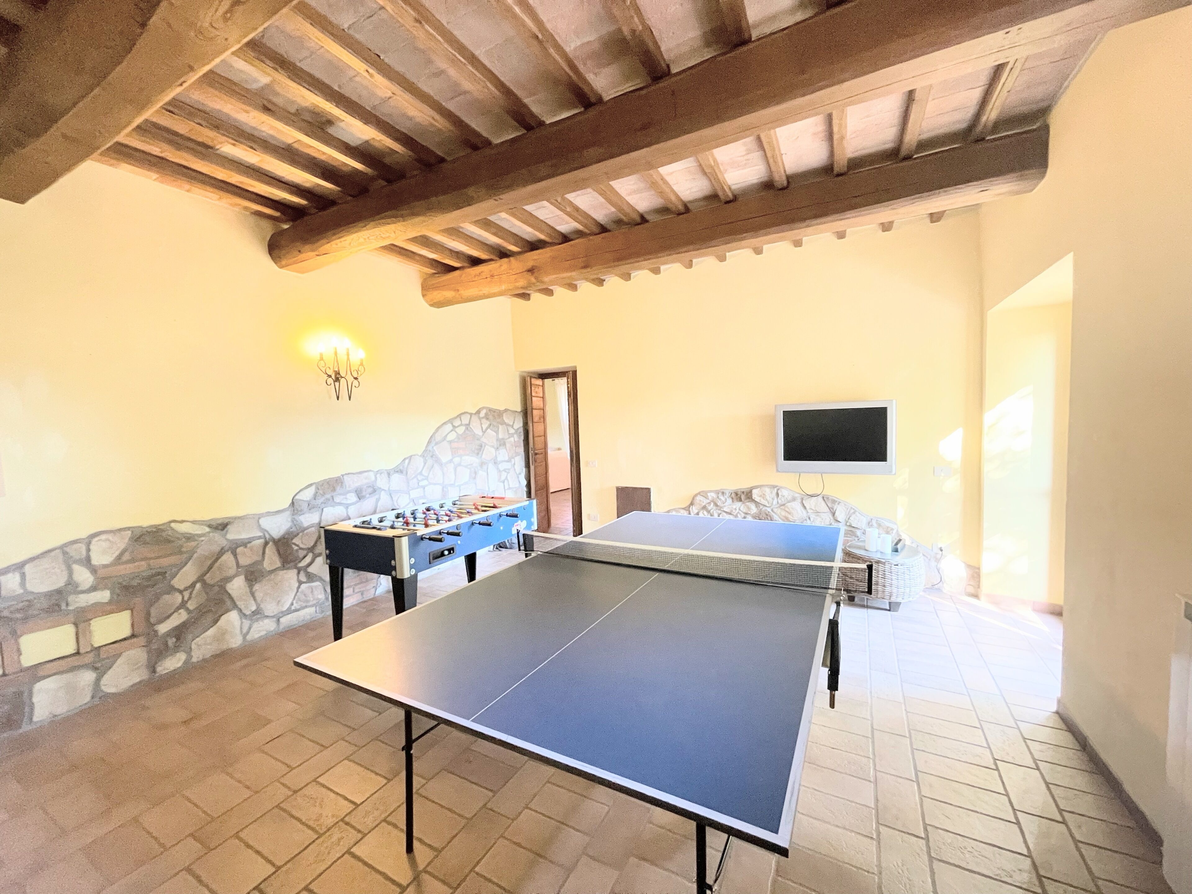 A room with a ping pong table, foosball table, stone wall, and a TV mounted on the wall.