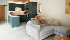 A modern kitchen with dark green cabinets, a white countertop, and a built-in oven. A light grey sectional sofa with decorative pillows is in the living area, and a round wooden table is visible.