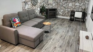 A living room with a stone wall, a gray sofa, a small round table, and a fireplace. - Adorable 1 Bedroom Suite With Kitchen & (Cambridge)