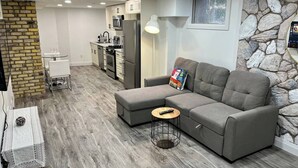 A modern living room with a grey sectional sofa, a small round coffee table, and a kitchen area with white cabinets and stainless steel appliances. - Adorable 1 Bedroom Suite With Kitchen & (Cambridge)
