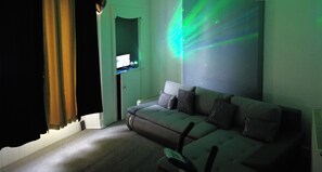Una sala de estar con sofá cama doble, televisor sobre un mueble bajo y pared con proyección de luz.