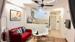 A compact living space with a red sofa, white chairs, a ceiling fan, a kitchen area, and a TV mounted on the wall. - Wind Chimes Hideaway - Apartment In Central Hanoi (Hanoi)