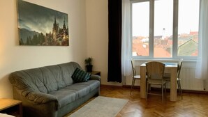 Ein Wohnzimmer mit grauem Sofa, einem kleinen Tisch mit Pflanze, einem Fenster mit Blick auf Gebäude und einem Gemälde eines Schlosses.