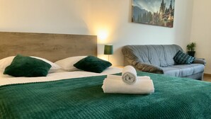 Ein ordentlich gemachtes Bett mit einer grünen Decke und einem zusammengerollten Handtuch, ein graues Sofa mit einem gemusterten Kissen und ein Gemälde eines Schlosses an der Wand.
