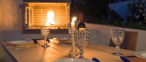 An outdoor dining table with plates, cutlery, and a candle, set against a backdrop of a lit fire.