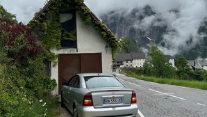 A silver car parked in front of a house with a garage, surrounded by greenery and mountains in the background. - Apartma Pohar- A Bit Different! (Bovec)