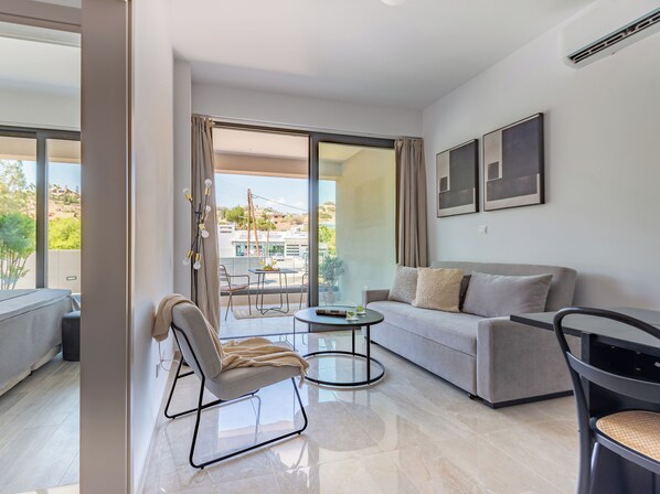 A modern living room with a grey sofa, a round coffee table, and a chair. There is a balcony with a view of the city and greenery.