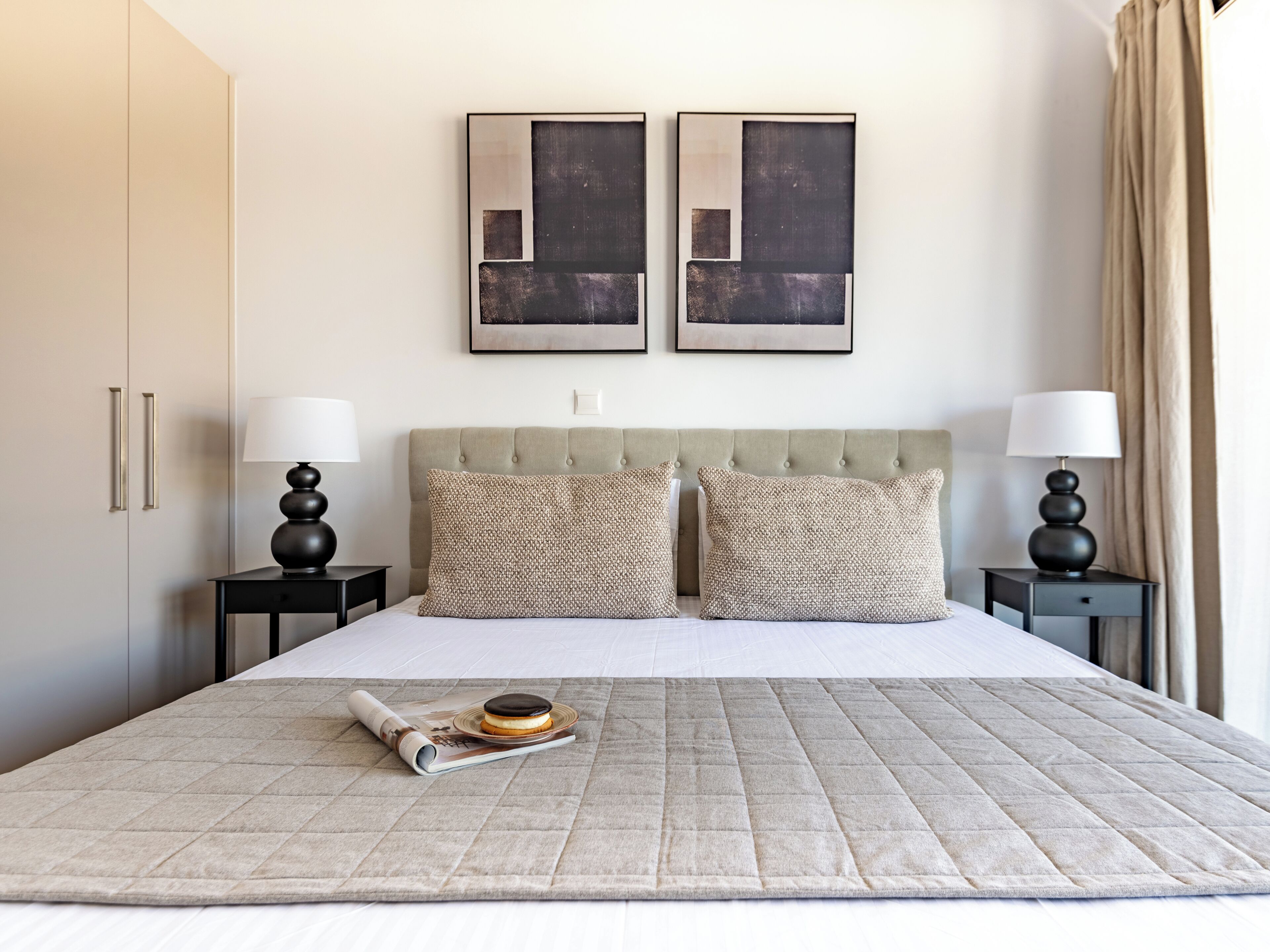 A neatly made bed with a quilted cover, two bedside tables with lamps, a headboard, and two framed abstract paintings on the wall.