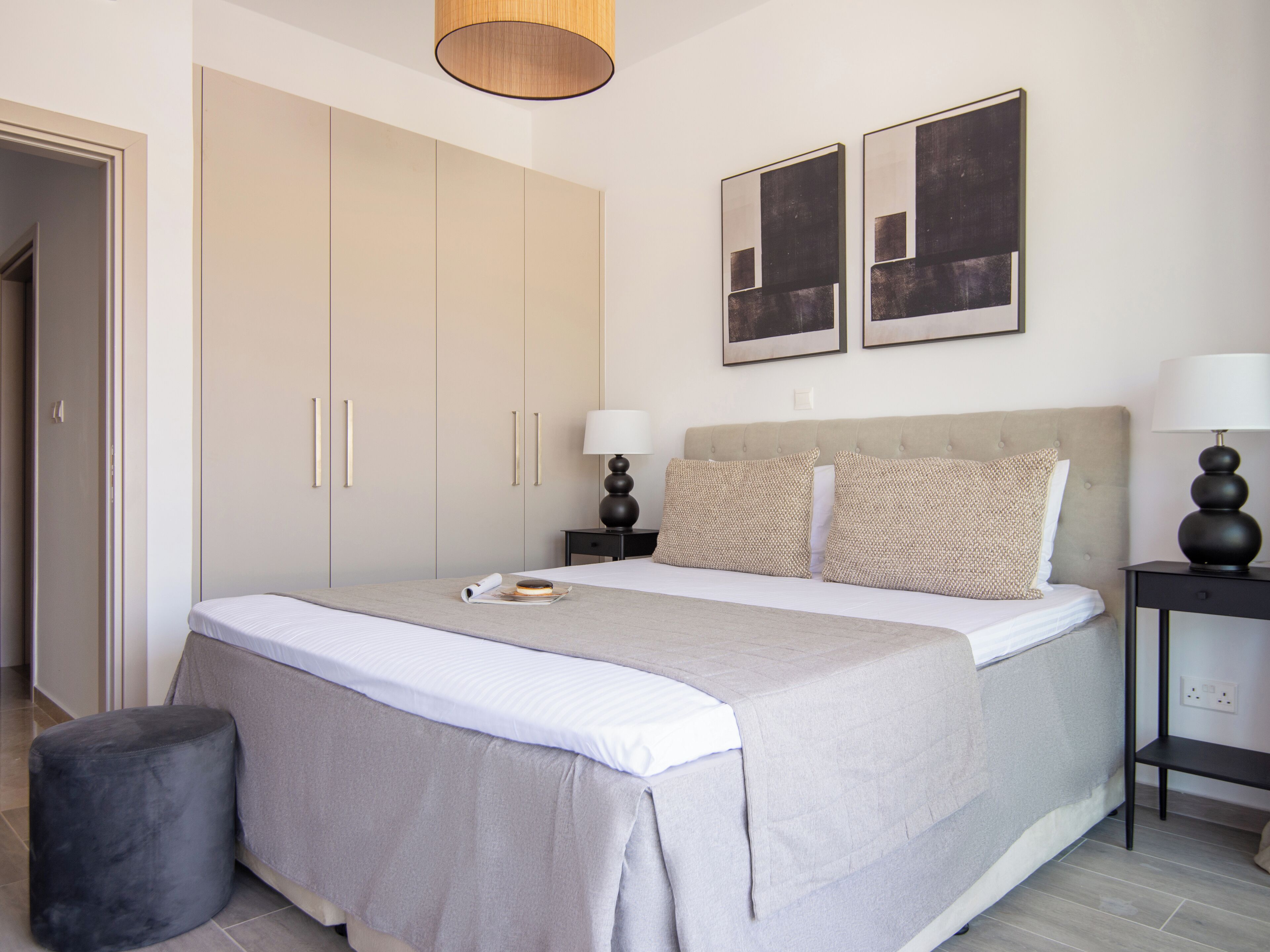 A modern bedroom with a large bed, a wardrobe, two bedside tables with lamps, and two abstract paintings on the wall.