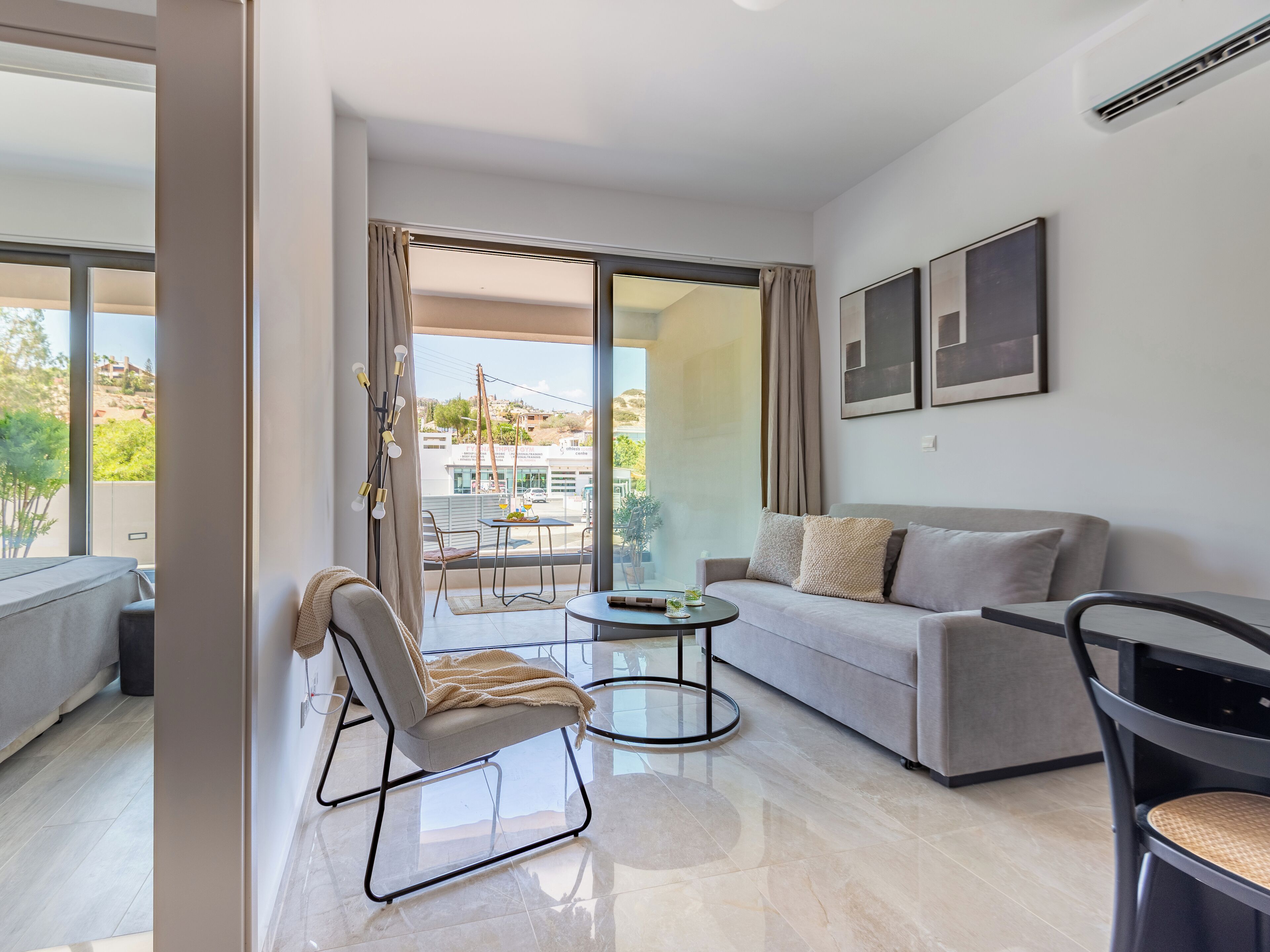 A modern living room with a grey sofa, a round coffee table, and a chair. There is a balcony with a view of the city and greenery.