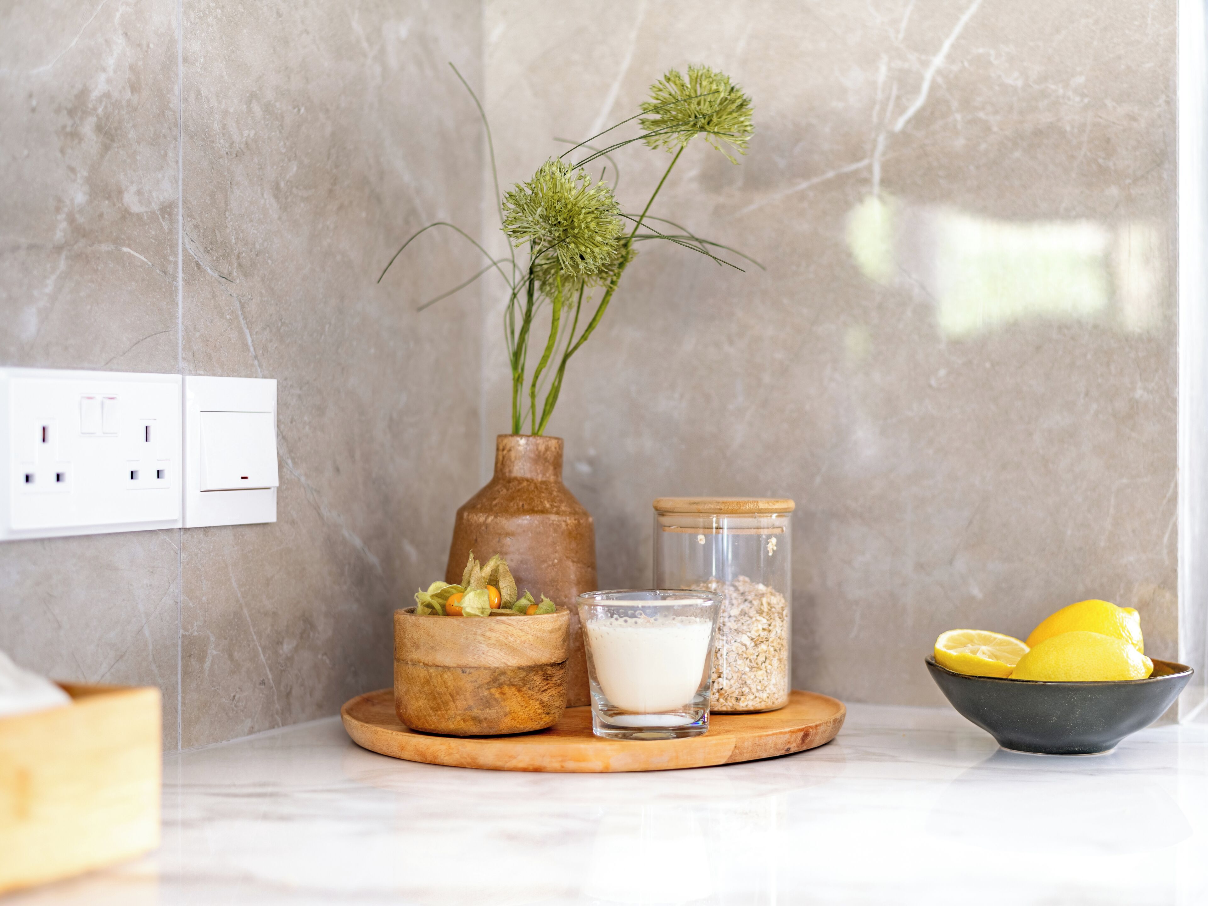 A kitchen counter with a vase of green plants, a bowl of lemons, a glass of milk, and a jar of oats.