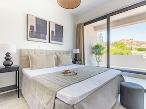 A modern bedroom with a large bed, two bedside tables, a window with a view of a mountainous landscape, and two framed pictures on the wall.