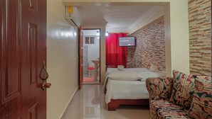 A hotel room with a wooden door, a bed with white bedding, a patterned sofa, a television on the wall, and a bathroom visible through an open door. - Room-single Room Near SDQ Airport 300 M (Santo Domingo Este)