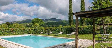 Une piscine rectangulaire bordée de pierres, entourée de pelouse et d’une haie. On y trouve des chaises longues et une pergola avec une table et des chaises.