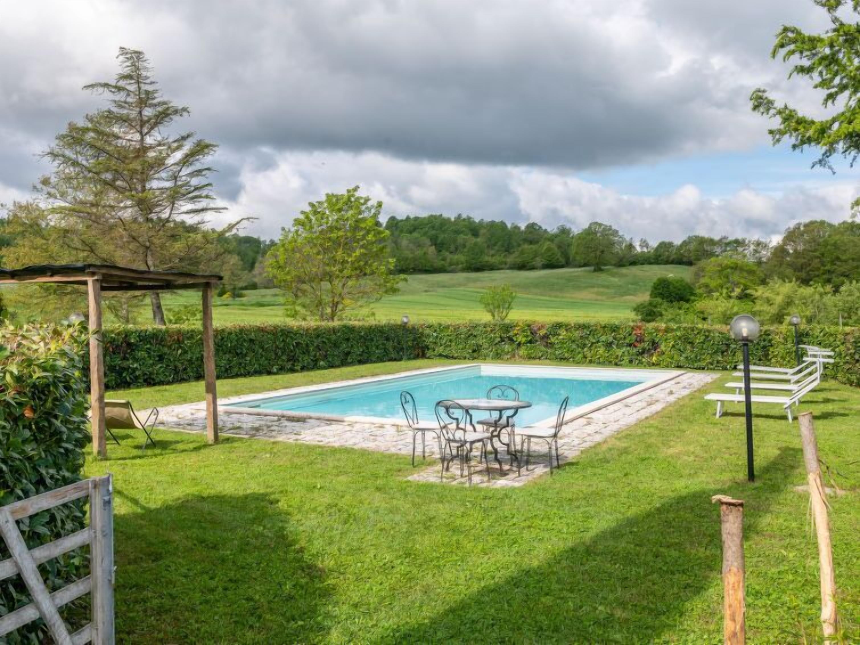 A rectangular pool with a surrounding stone deck, a wooden pergola, and a table with chairs.