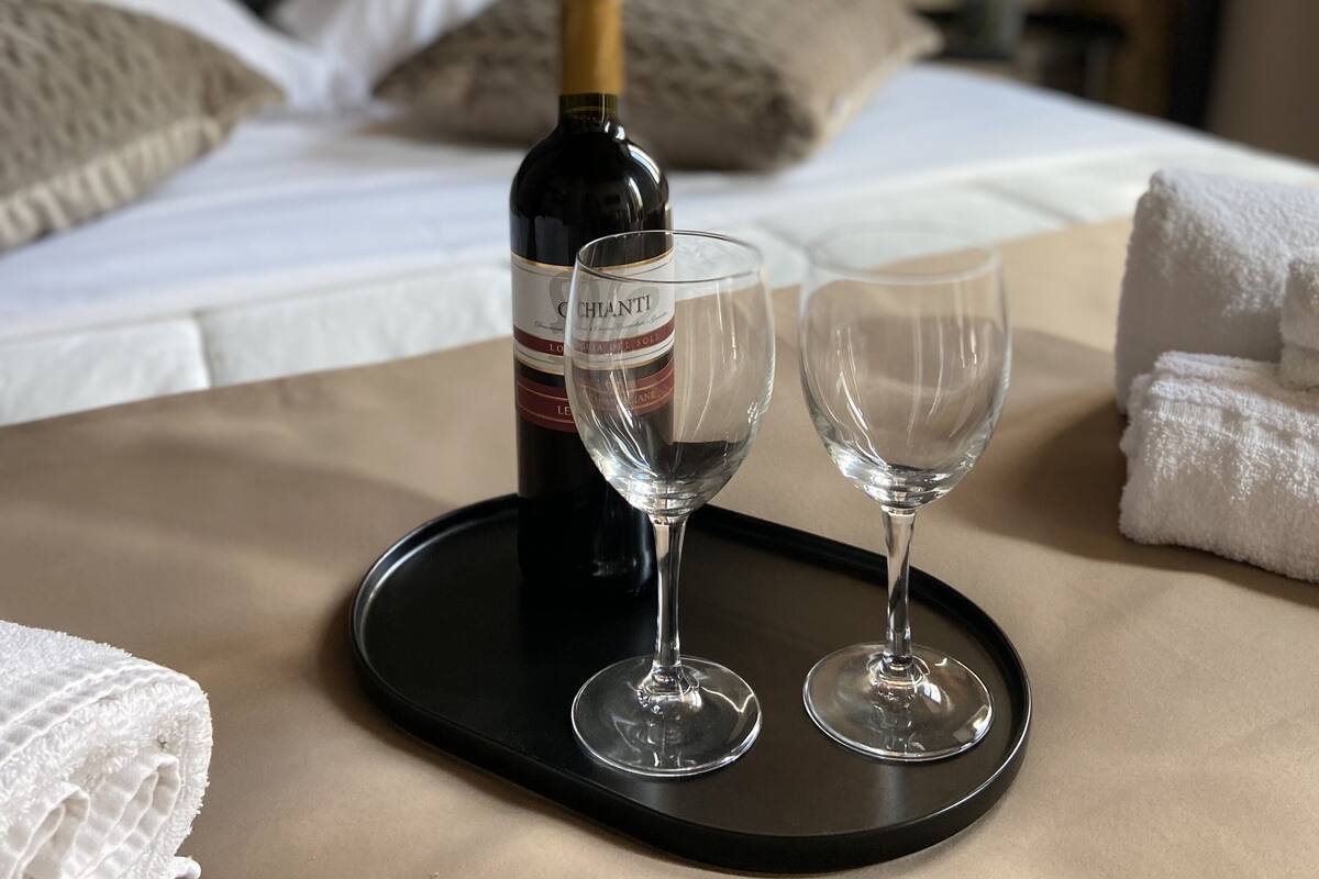 A bed with white linens, a bottle of wine, two empty wine glasses, and a tray on a table.
