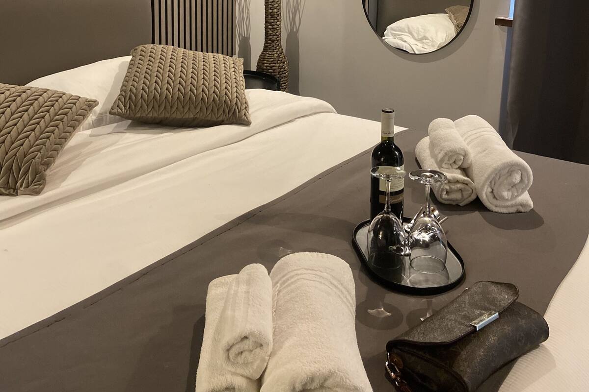 A modern hotel room with a neatly made bed, a bedside table with a wine bottle, glasses, and towels, a wall-mounted mirror, and a vertical light fixture.