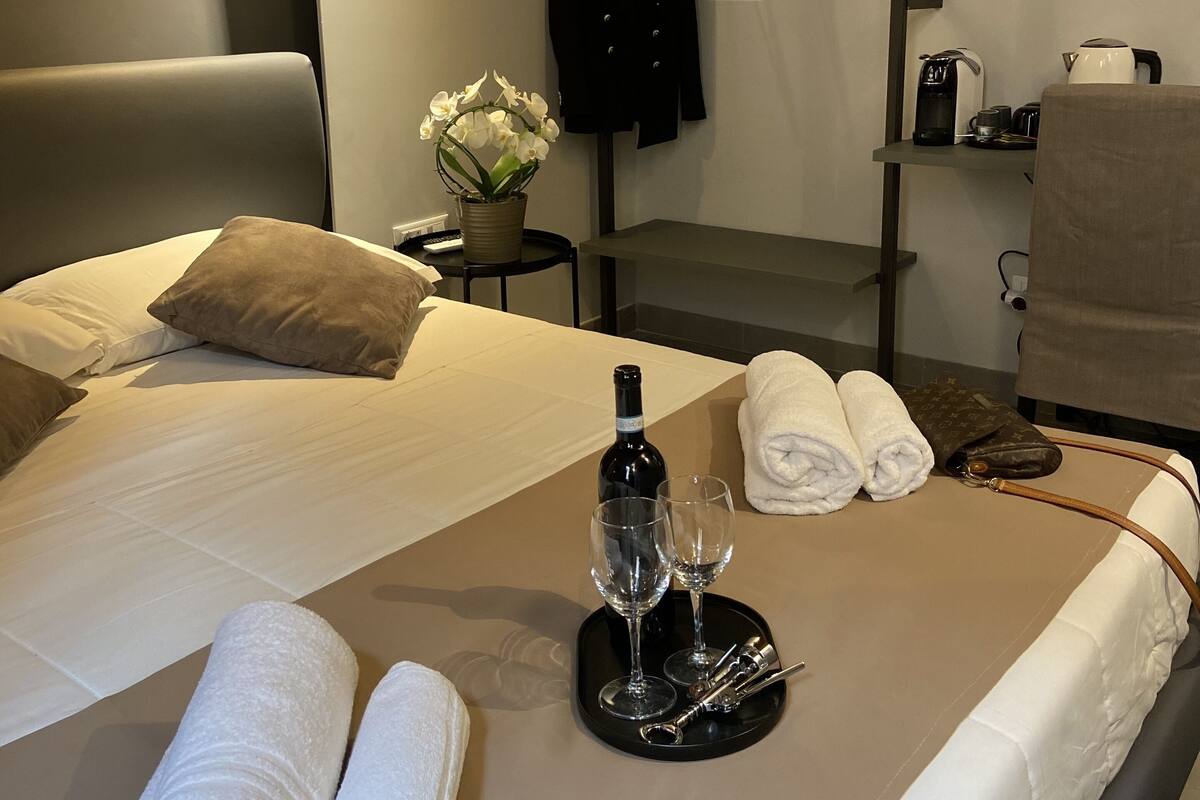 A hotel room with a bed, a side table with a bottle, glasses, and towels, a shelf with a kettle and a jacket, and a hanging coat rack.