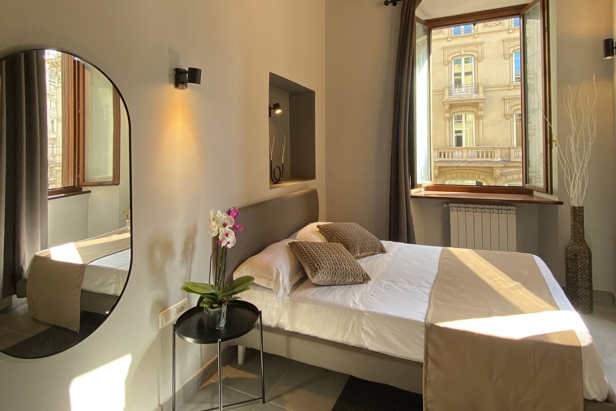 A bedroom with a large bed, a bedside table with a vase of flowers, a mirror, and a window with a view of buildings.