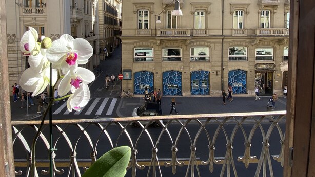 Vista de una calle con edificios, incluyendo uno con letreros de 'For Lease', y un balcón con una planta.