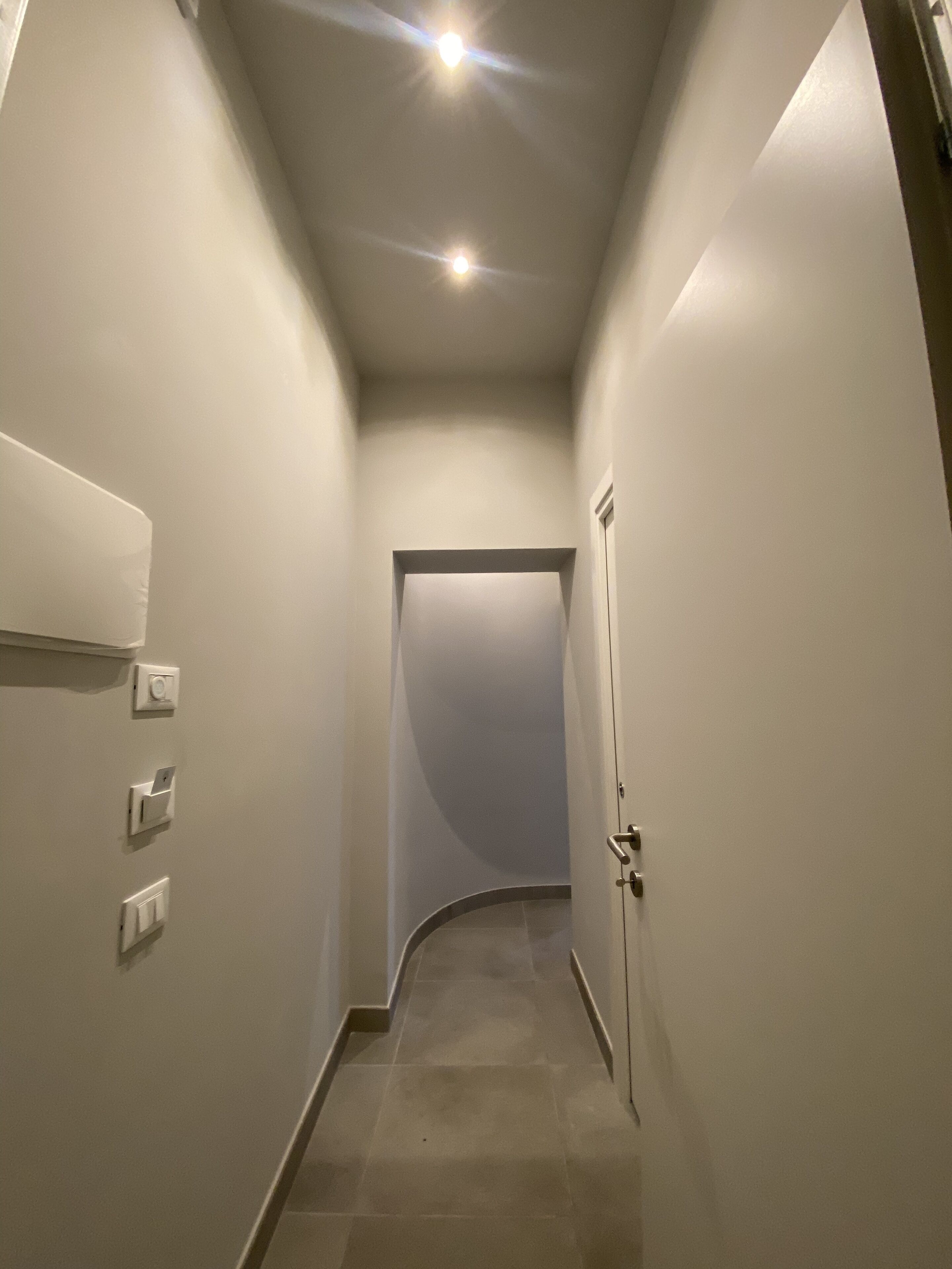 A narrow hallway with a door at the end, recessed lighting, and three wall-mounted light switches.