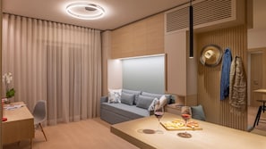 A modern, well-lit room with a sofa, a dining table with a wine glass and cheese board, and a desk with a plant.