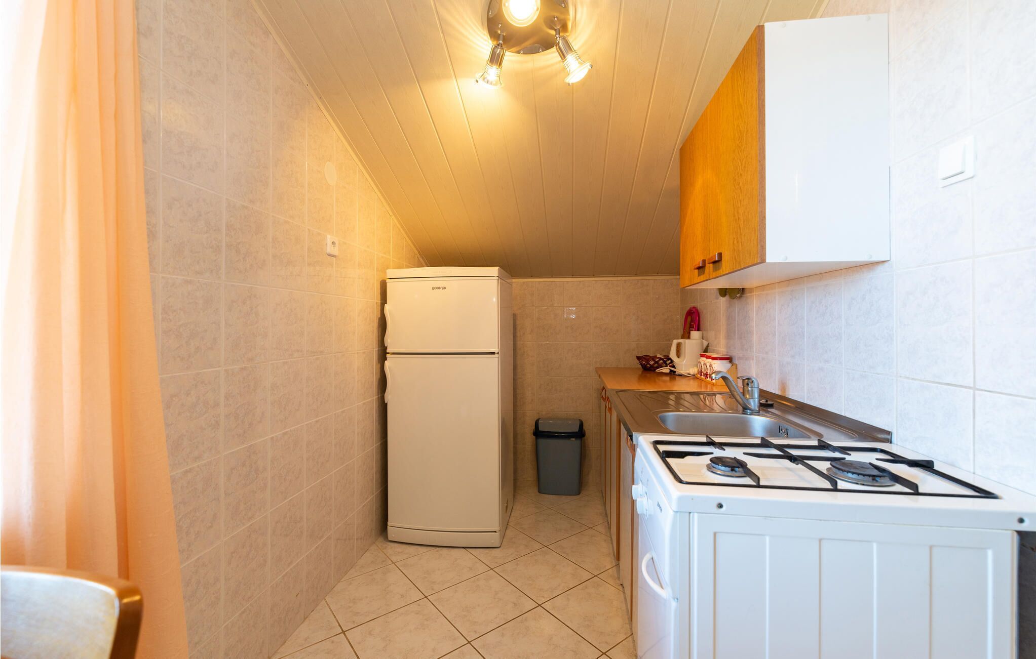 A compact kitchen with a refrigerator, stove, sink, and tiled walls.