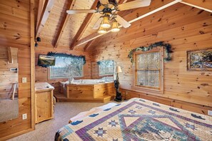 A cozy cabin room with wooden walls, a ceiling fan, a large bed, a desk, a window with blinds, and a fireplace. - Large 5 BR w/ Hot Tub, Mtn View, Pool Table, WiFi + Close to Pigeon Forge Strip (Sevierville)
