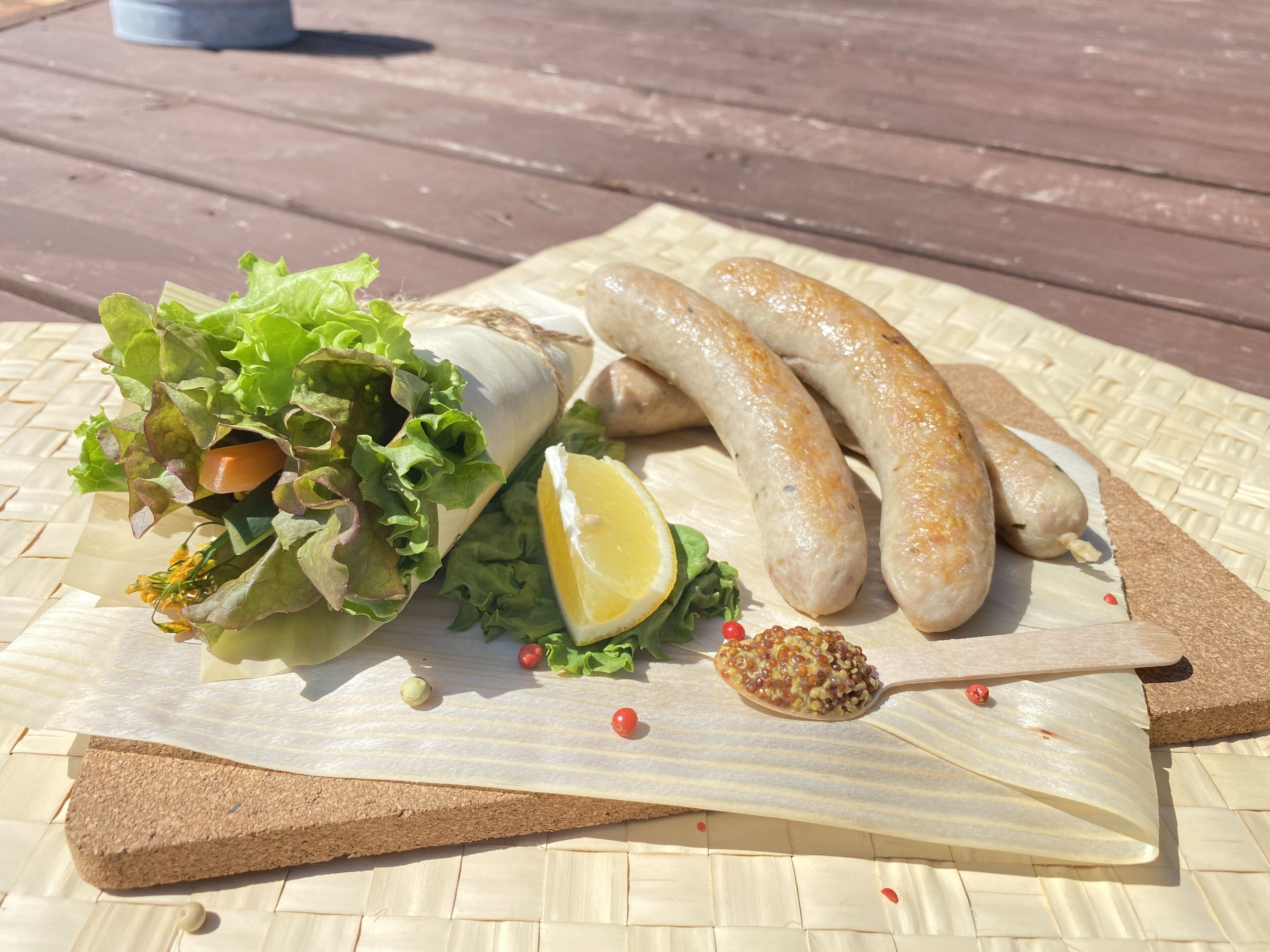 Et træbord med en vævet måtte, en træskærebræt og en kurv med friske grøntsager, herunder salat, en citron og kirsebær tomater.