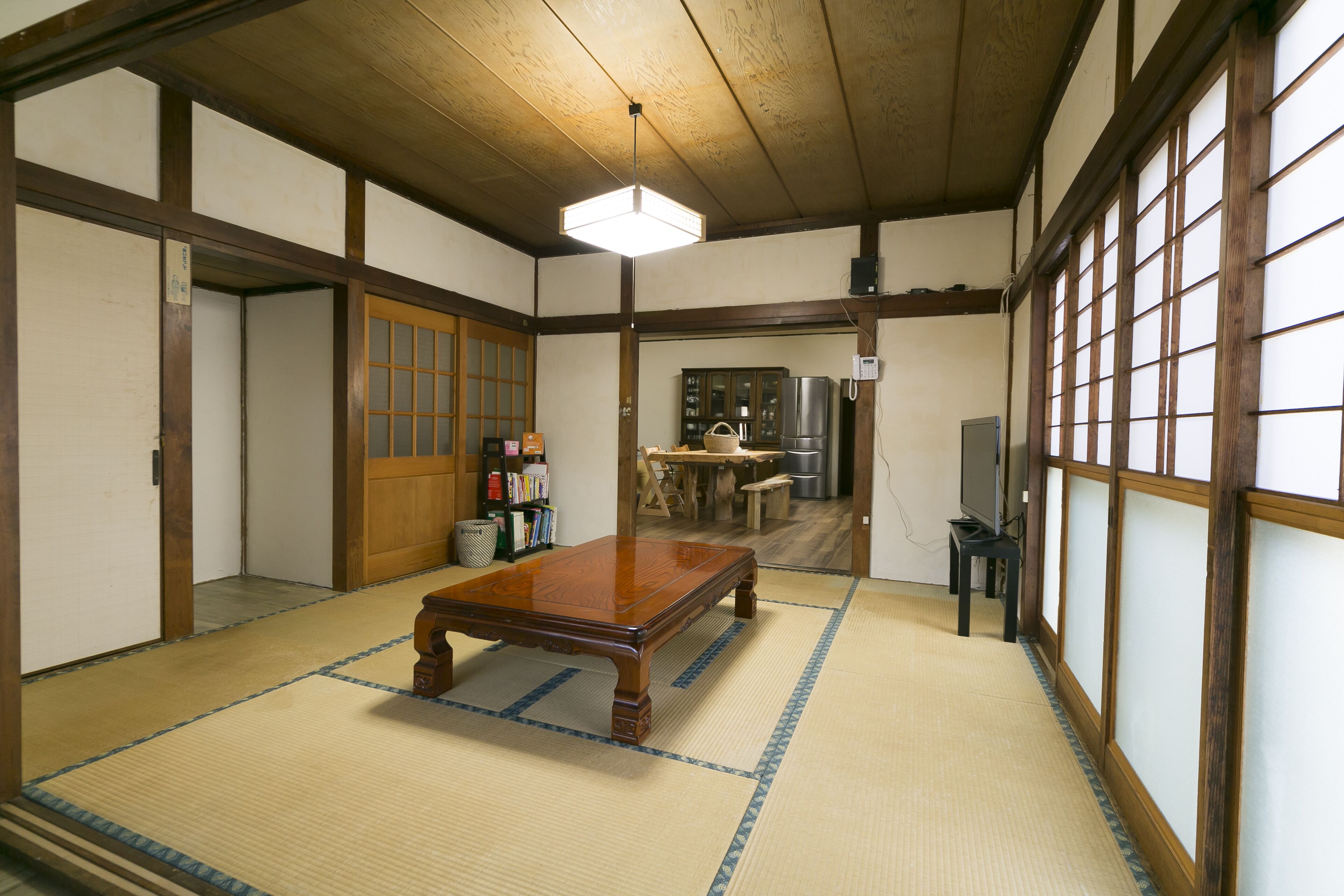 A traditional Japanese room with tatami flooring, a wooden table, a television, and a kitchen area in the background.
