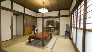 A traditional Japanese room with tatami flooring, a wooden table, a television, and a kitchen area in the background.
