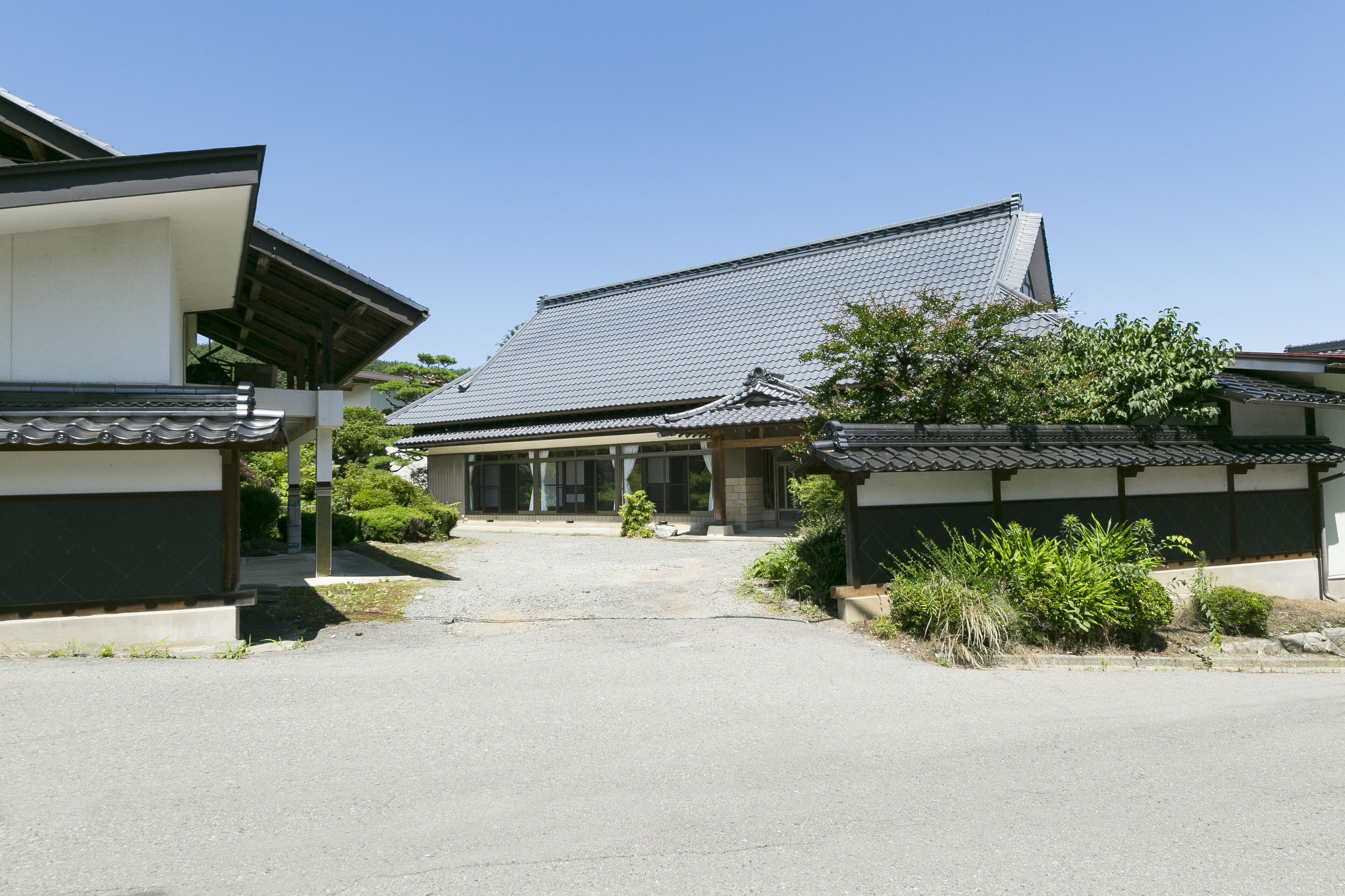 A traditional Japanese building with a tiled roof, surrounded by greenery and a gravel driveway.