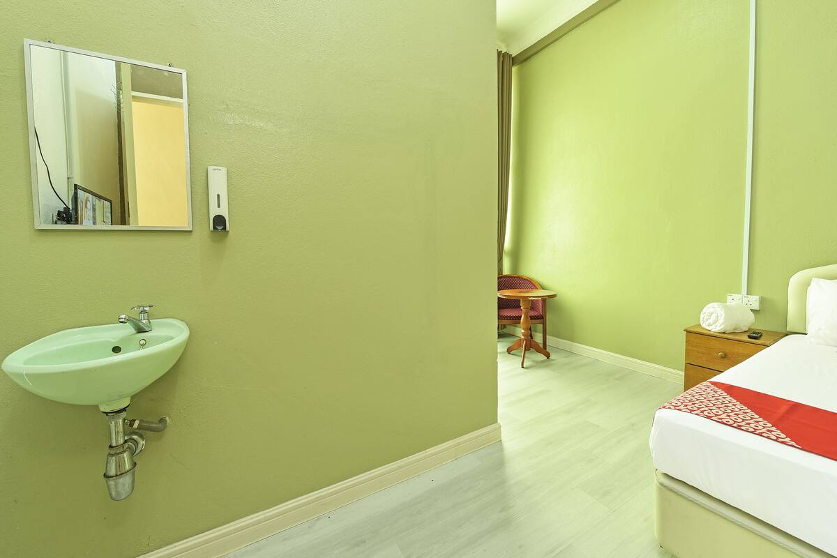 A hotel room with a green wall, a white bed with a red and white blanket, a wooden nightstand, a small wooden table, and a green sink with a mirror above it.