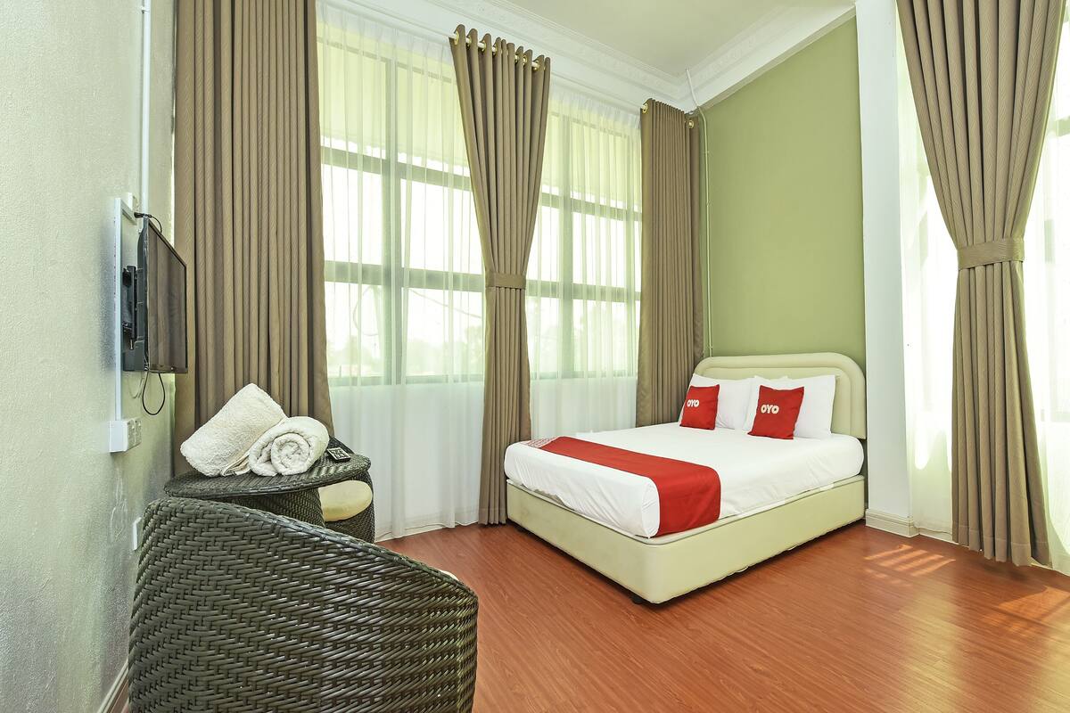 A hotel room with a bed, a television mounted on the wall, a wicker chair, and curtains.