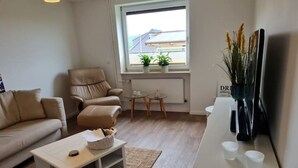 A modern living room with a sofa, a recliner, a coffee table, and a small side table. There is a window with a view of a building outside. - Schöne Ruhige Ferienwohnung mit Terrasse (Flensburg)