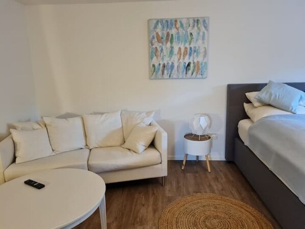 Ett modernt rum med en beige soffa, ett litet runt bord, ett sängbord med en lampa och en säng med kuddar.