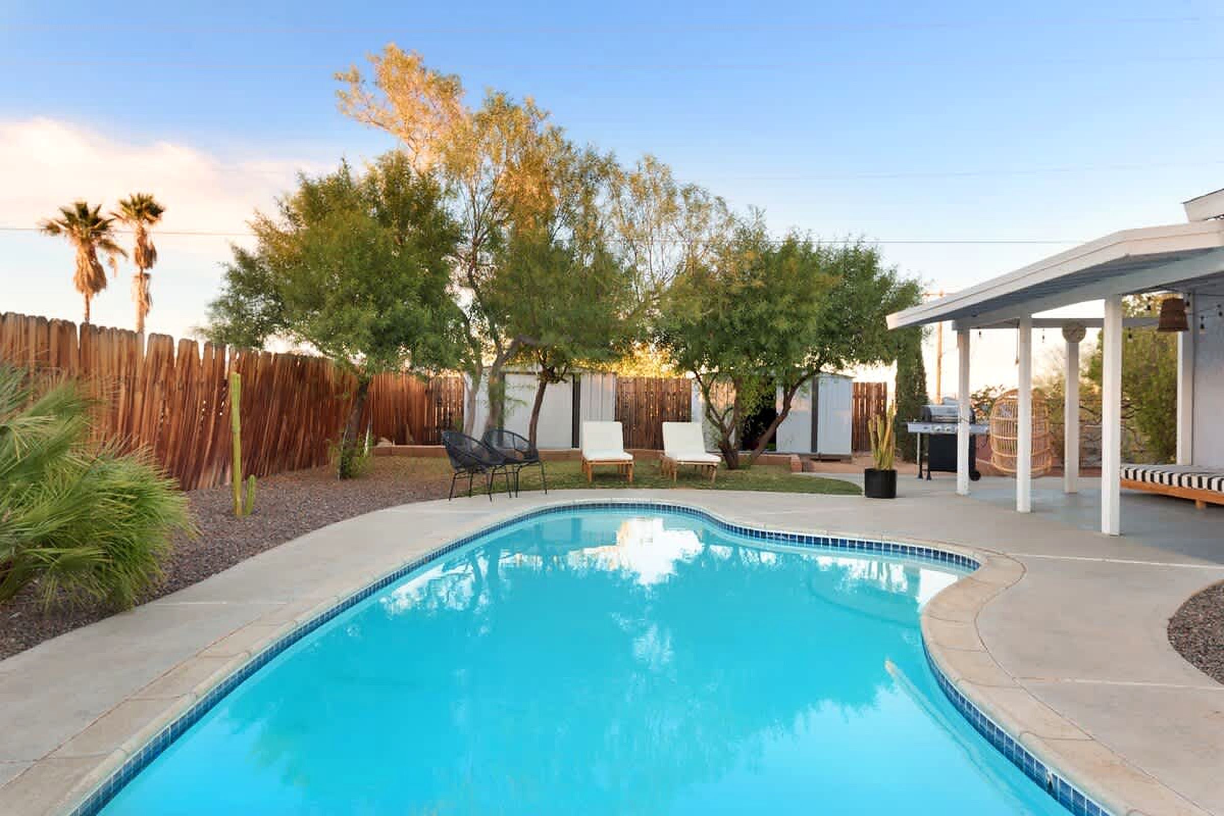 A swimming pool with a wooden fence, lounge chairs, and a covered patio area.