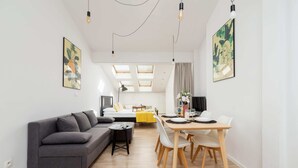 A modern living room with a grey sofa, a dining table with chairs, and a small side table. There are skylights above the dining area and a bed in the background.
